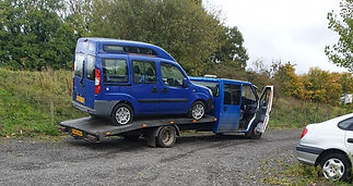 A blue van is on a flatbed tow truck beside a rural road