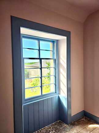 A window with blue frames looking out onto a green landscape, with light pink walls and wooden floor
