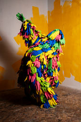 A figure stands with one arm pointed up, bent precariously backwards. It is covered by a ghillie suit made of gloves in pink, blue, green and yellow.