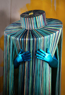 A wide, flat brimmed hat made of floor-length pieces of string in various shades of blues. A pair of blue elbow length gloves protrude from the hat, but the wearer cannot be seen.