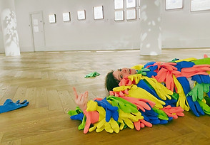 Naoibh lays on wood floor. They are wearing a suit made of colourful gloves and making a silly face at the camera.