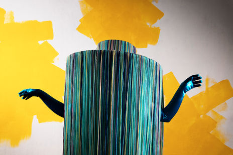 A wide, flat brimmed hat made of floor-length pieces of string in various shades of blues. A pair of blue elbow length gloves protrude from the hat, but the wearer cannot be seen.