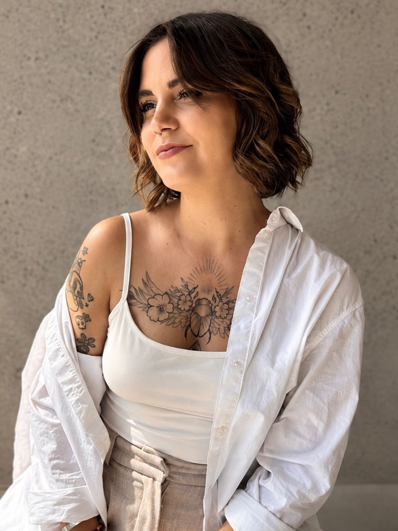 Portrait d'Anouck, chemise blanche ouverte sur un débardeur, le regard doux tourné vers le côté, devant un mur gris clair