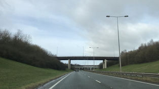 Empty highway under cloudy sky, with overpasses, streetlights, grassy embankments, and a few cars ahead