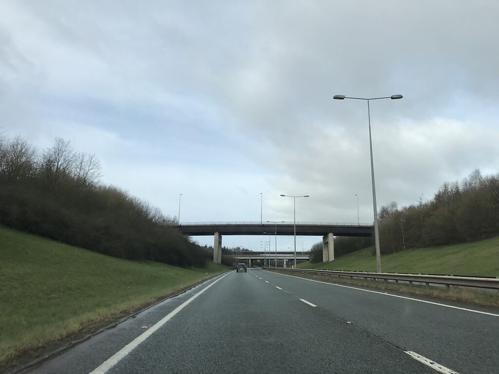 Empty highway under cloudy sky, with overpasses, streetlights, grassy embankments, and a few cars ahead