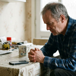 Man in blue plaid shirt sits pensively at a kitchen table with a smartphone, newspaper, and floral mug. Soft floral wallpaper in background.