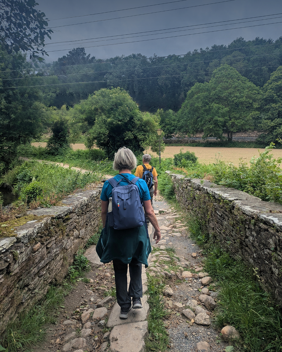 Two pilgrims crossing a stone bridge on the Camino de Santiago near Sarria, with green Galician countryside under a stormy sky