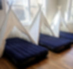 Three blue air mattresses under white teepee tents in a sunlit room with wooden floors. Large windows show bare trees outside.