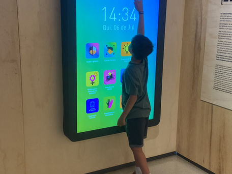 Criança utilizando uma grande tela interativa em formato de smartphone dentro do SESI Lab, em Brasília. A instalação digital exibe aplicativos e conteúdos educativos relacionados à tecnologia, enquanto o visitante interage com a exposição.