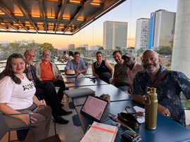 Grupo de pesquisadores, professores, comunicadores científicos e representantes de instituições reunidos em mesas no terraço do SESI Lab, em Brasília, durante encontro sobre a criação do Museu de Ciência e Tecnologia de Brasília. Ao fundo aparecem prédios da região central da capital ao entardecer.