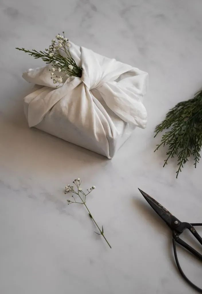 pacote de presente embrulhado com um tecido branco e efeitado com um ramo de folhas verdes e pequenas flores brancas