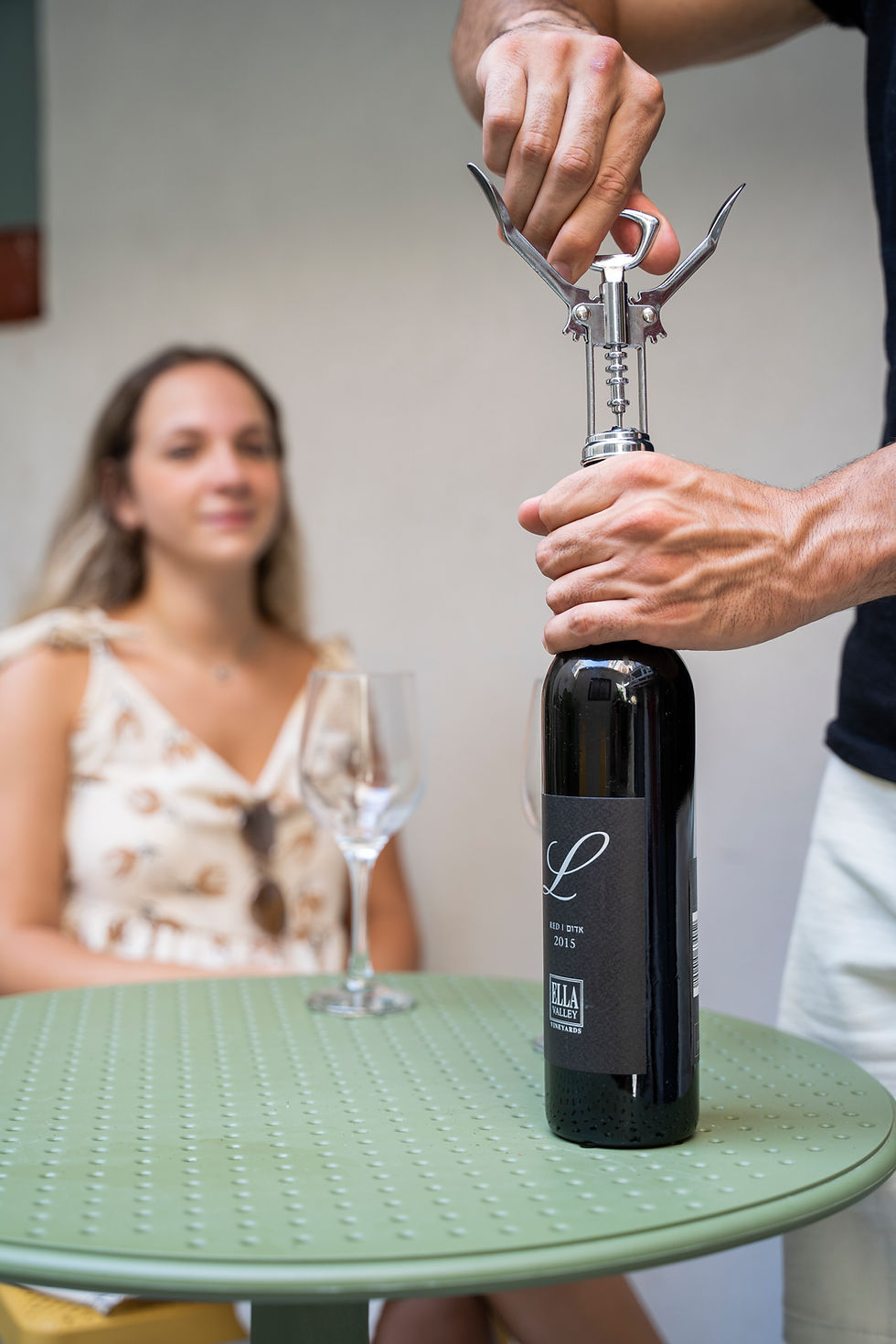 Person opening wine bottle with corkscrew, woman in background