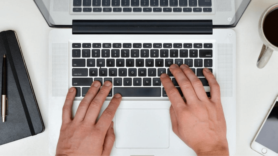 Hands type on a laptop computer. On the sides of the computer are a notebook and pen, phone and a coffee