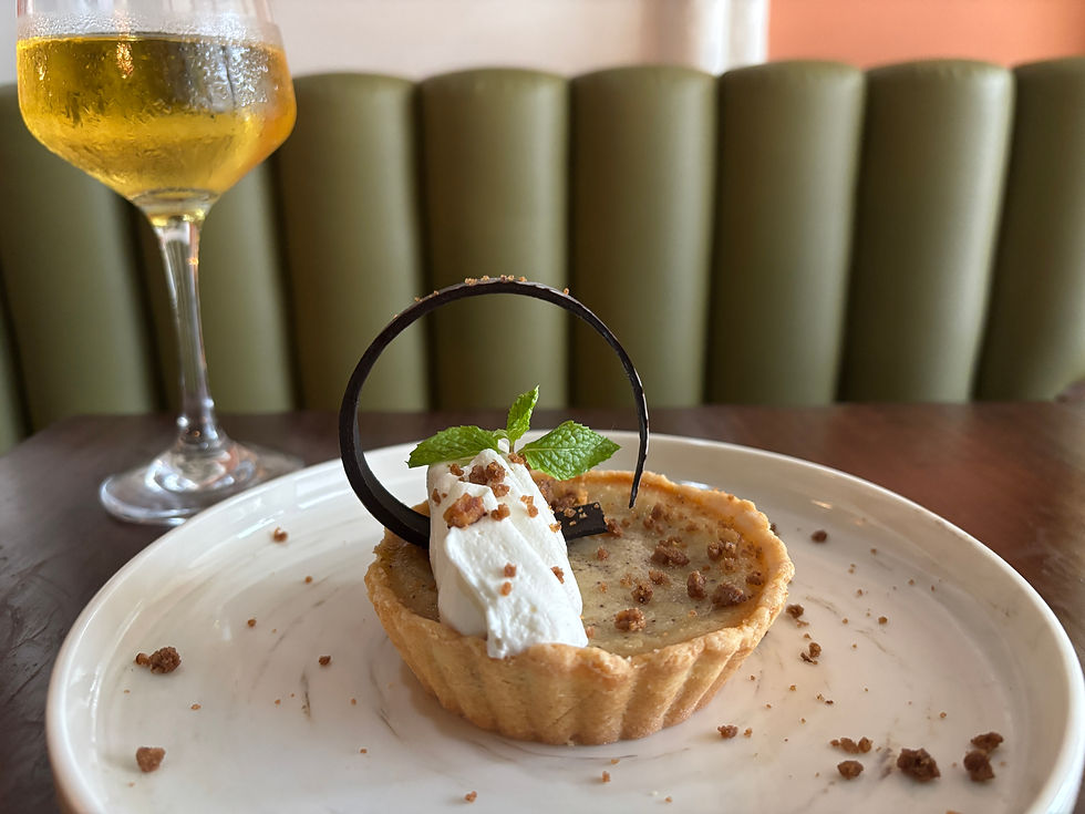 A tart topped with whipped cream, a chocolate curl, and mint on a white plate. A glass of golden drink sits nearby, on a wooden table.