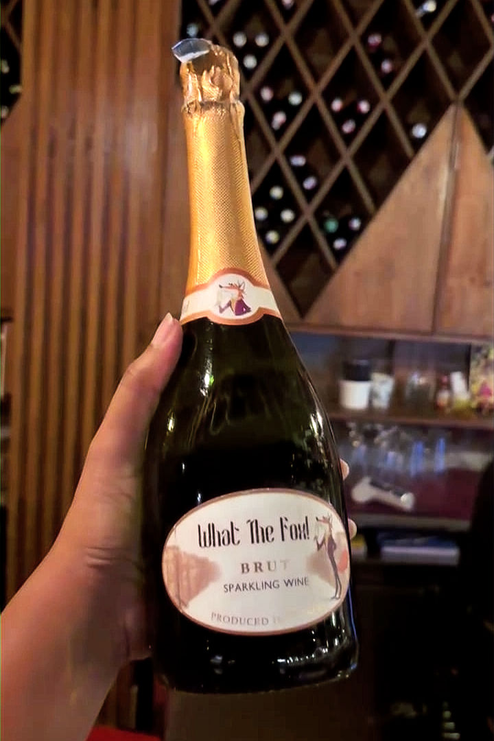 Hand holding a What The Fox? Brut sparkling wine bottle in a warm wine cellar with wooden racks.