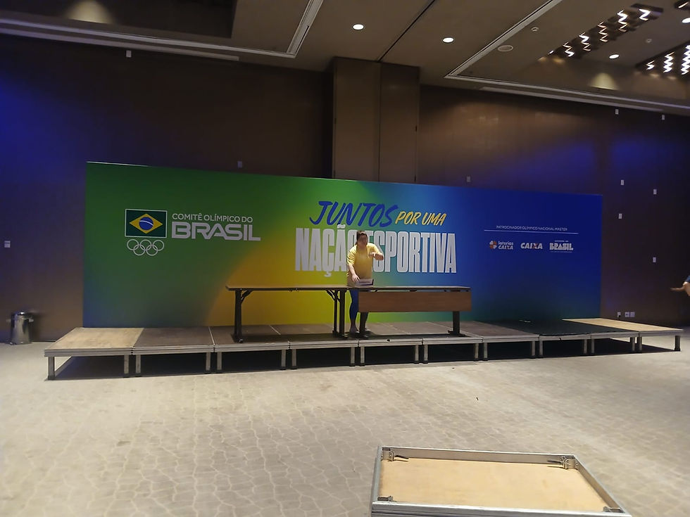 Mulher em palco indoor ajusta mesa diante de banner verde-azul com texto Juntos por uma Nação Esportiva e Comitê Olímpico do Brasil