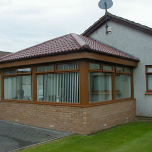 A brick and wood conservatory with large windows and vertical blinds attached to a suburban house.