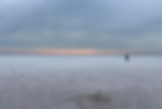 The vast white salt flats look like a snow-covered plain. One man is seen in the distance amid a rising sun covered by looming clounds.