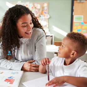 Female infant school teacher working one on one with a young schoolboy, sitting at a table smiling at each other, close up