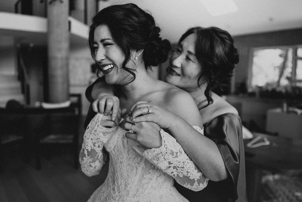 Mother helping bride into lace wedding gown during emotional getting-ready moment.
