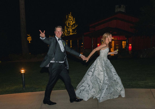 Bride and groom running hand in hand at night outside reception venue under string lights.