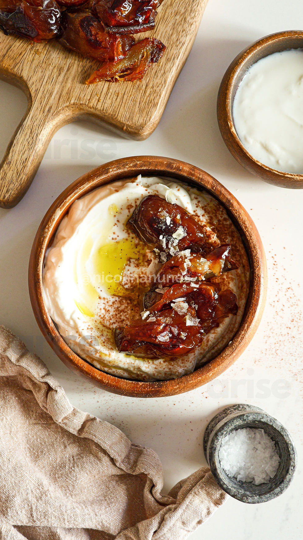 Wooden bowl with creamy yogurt, roasted dates, olive oil, and salt flakes on a pale surface. Rustic vibe with a fabric napkin.