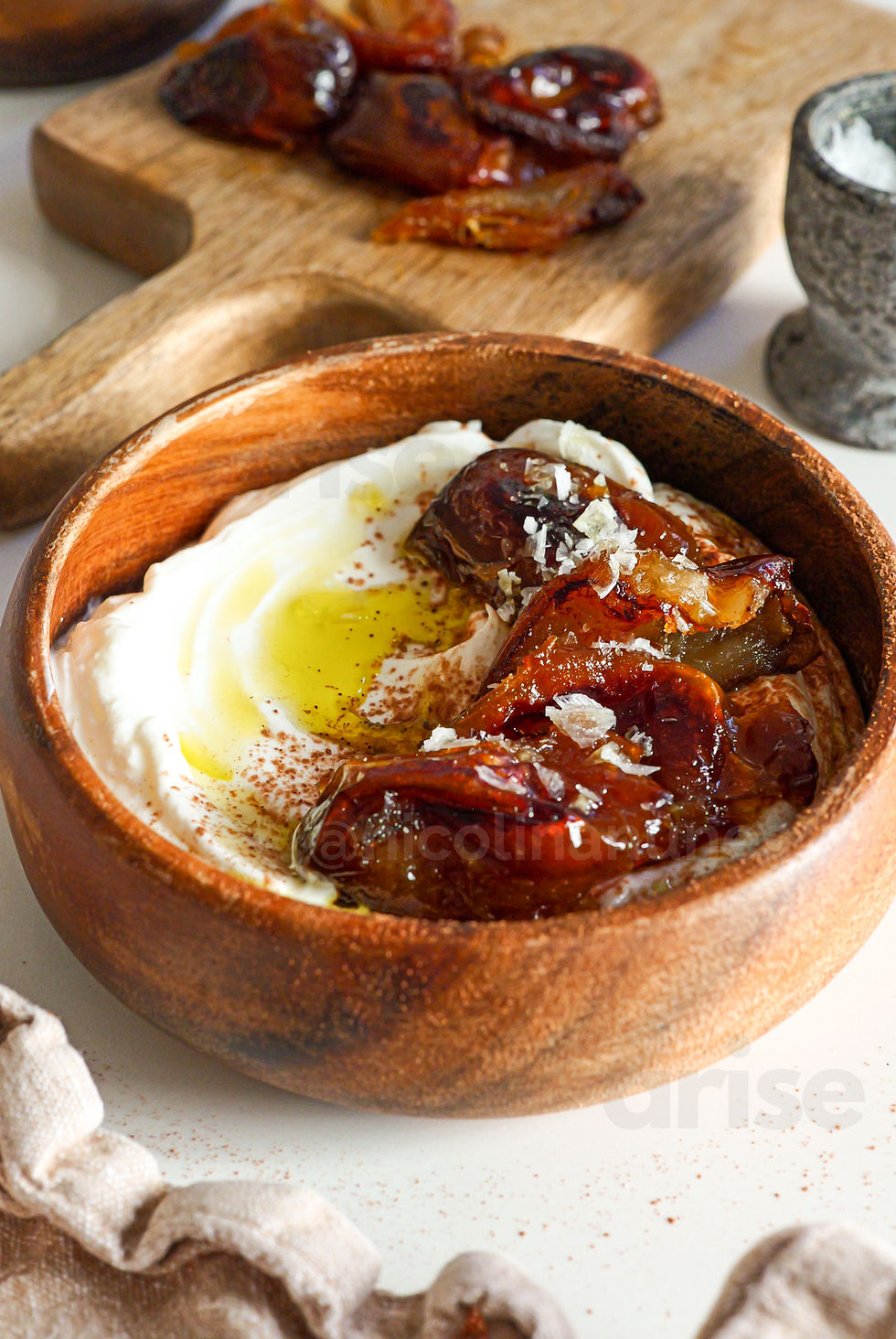 Wooden bowl with yogurt, roasted figs, and olive oil, garnished with salt on top. Background: cutting board with more figs.