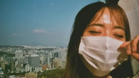 Woman wearing a mask with eyes closed, enjoying the sunlight. Background shows a cityscape under a clear blue sky. Calm mood.