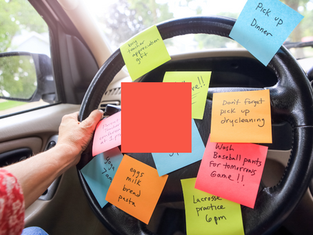 A car steering wheel covered with colorful sticky notes, each listing various tasks and reminders like 'Pick up dinner,' 'Yoga 7 am,' and 'Parent-teacher conference at 1 pm.' A hand grips the wheel, symbolizing a busy, multitasking lifestyle. A red square obscures part of the wheel, and the car's interior and blurred outdoor greenery are visible in the background.