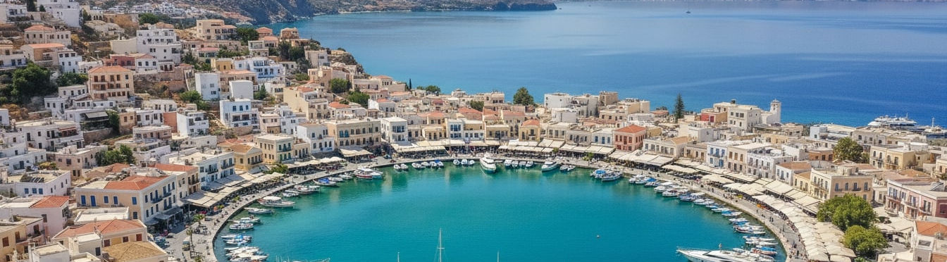 Panoramic view of Agios Nikolaos harbor town in Crete showing Lake Voulismeni surrounded by colorful Mediterranean buildings, traditional fishing boats in the marina, and turquoise waters of the Gulf of Mirabello with mountains in the background