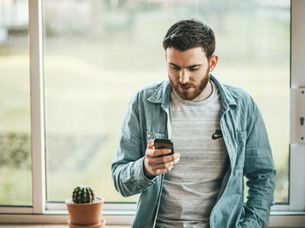 Ein Mann steht vor einem Fenster angelehnt an einen Tisch und blickt nach unten auf sein Smartphone