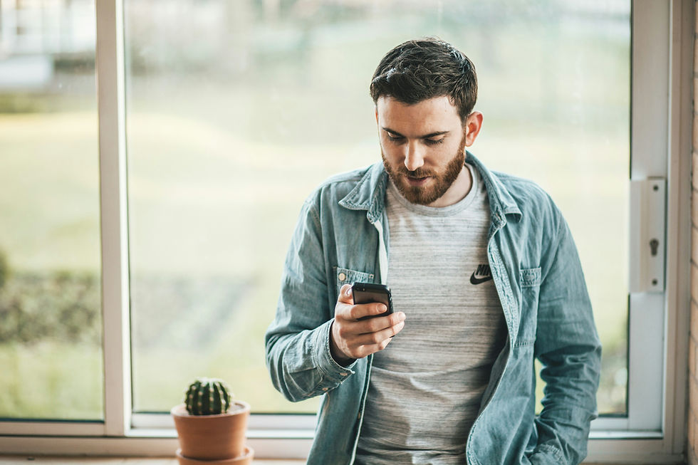 Ein Mann steht angelehnt an einen Tisch vor einem Fenster und sieht nach unten auf sein Smartphone