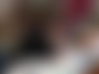 Harley leans over a piece of fabric, smiling at the camera. He is in a workshop with arts and crafts materials on the table around him, as well as on shelves in the background.