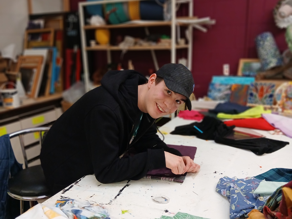 Harley leans over a piece of fabric, smiling at the camera. He is in a workshop with arts and crafts materials on the table around him, as well as on shelves in the background.