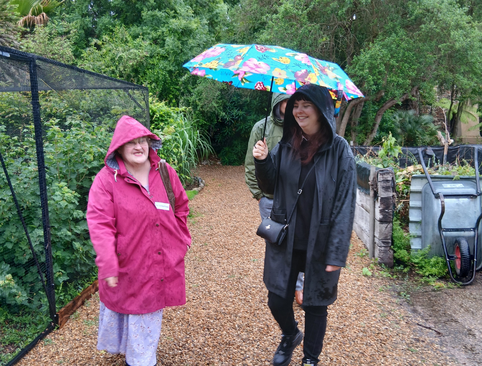 Members and staff walk to the Rt Revd Graham Usher's lunch through a garden with trees and plants on either side. Two people at the front are turned towards each other, smiling and talking as they shelter under umbrellas.