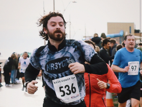Matthew Northcote runs the Norfolk Half Marathon, wearing a black and white tie dye top with the number 536 on his white marathon label. Behind him are other runners and supporters.