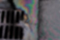 An oil slick against the backdrop of an asphalt road flows into a storm drain through a gr