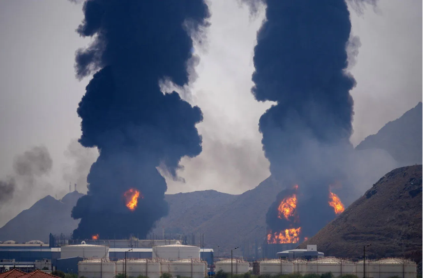 An industrial energy facility with storage tanks in the foreground as large fires burn on nearby hills, sending thick black smoke into the sky, illustrating damage to oil and gas infrastructure.