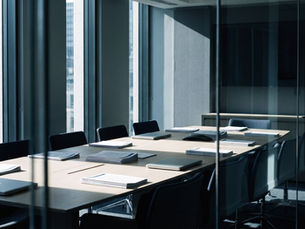 An empty boardroom seen through a glass wall, with a long pale wood table, closed laptops, stacks of printed documents and dark chairs, lit by daylight from tall city windows.