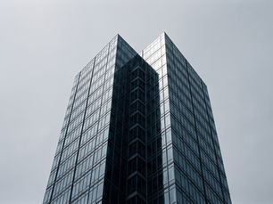A tall glass and steel office tower photographed from below against a flat grey sky, its two wings meeting at a sharp vertical edge.