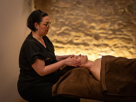 Esthéticienne en noir massant le visage d’une cliente allongée, dans un spa aux tons bruns et lumière douce.