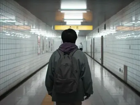 A movie still from Exit 8 (2025). The protagonist is standing in a clean, white tiled hallway. A bit further ahead, the sign Exit 8 is hanging from the ceiling. 