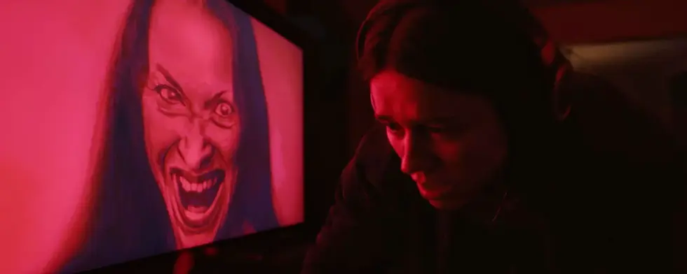 A movie still from Undertone (2026). Evy, the movie's main character, is in front of a TV screen, looking terrified. On the screen, in red hue, we can see a woman's distorted face, she looks insane.