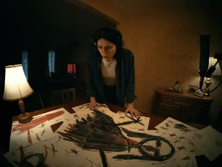 A movie still from Undertone (2026). Evy is standing above a table full of papers that when put together form a drawing of a scary woman-like shape.