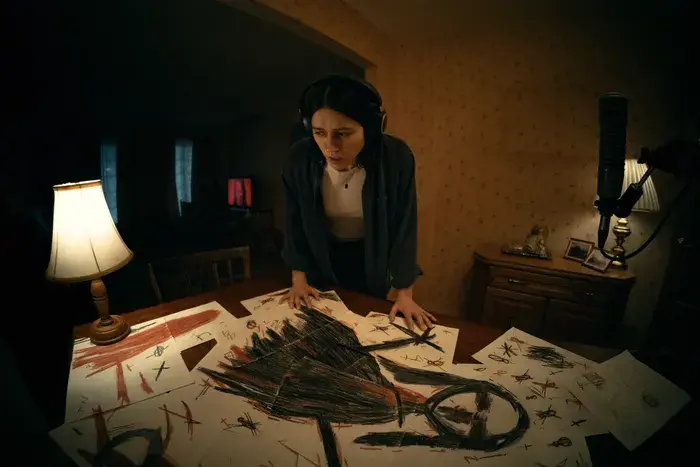 A movie still from Undertone (2026). Evy is standing above a table full of papers that when put together form a drawing of a scary woman-like shape.