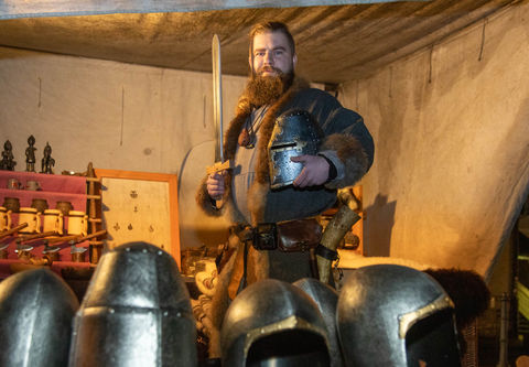 Ein junger, bärtiger Mann hält in einer Hand ein Schwert, in der anderen einen Helm an seinen Körper. Der Mann steht in seinem Marktzelt, vor ihm ist eine Reihe von mittelalterlichen Helmen zu sehen.
