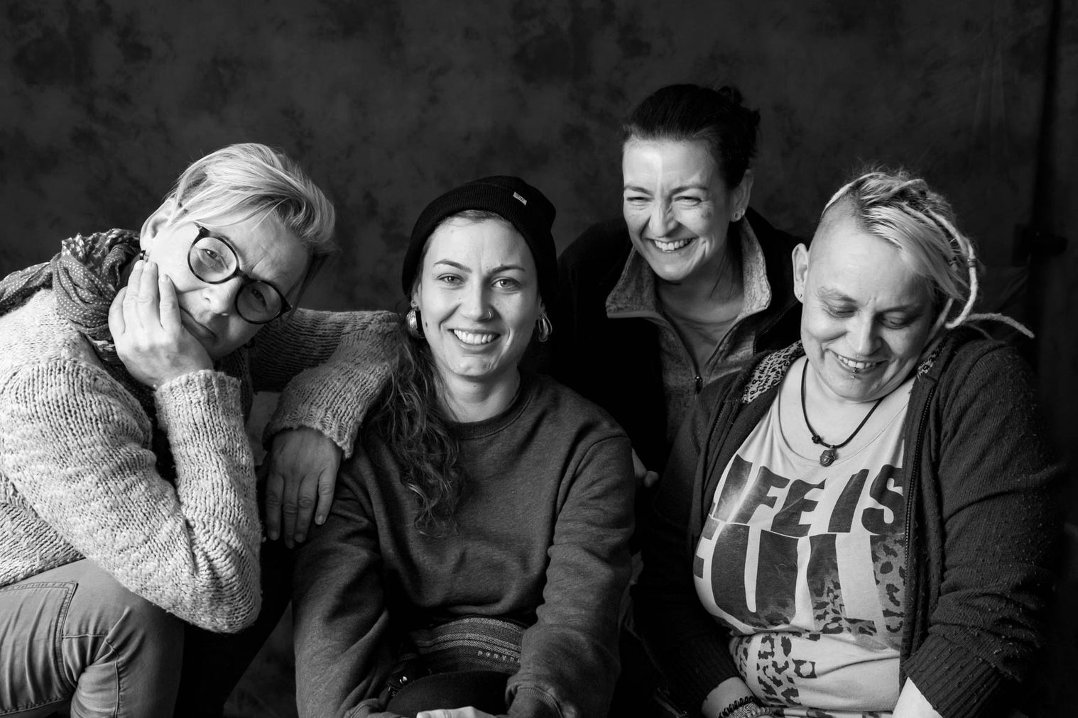 Schwarzweiß Fotografie von vier Frauen vor dunklem Hintergrund. Diese schauen in die Kamera und auch nicht. Die Frauen sind jung bis hin zum mittleren Alter und tragen helle und dunkle Kleidung. Offensichtlich fühlen sie sich zu viert wohl. Das Porträt wurde in der Vision e.V. in Köln-Kalk erstellt, einem Verein für innovative Drogenselbsthilfe.
