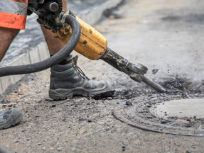 Ein Arbeiter entfernt mit einem Abbruchhammer, der in der Arbeitersprache "Stemm" genannt wird, sauber den Asphalt um den Bereich eines Kanaldeckels. Der Kanaldeckel ist ebenerdig mit dem umgebenden Asphalt verbunden.