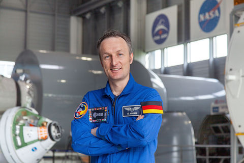 Astronaut Matthias Mauer steht im ESA-Ausbildungszentrum in Köln Porz-Wahn und hat die Arme verschränkt. Im Hintergrund sind Module der ISS und weitere Raumfahrt-Module zu erkennen.