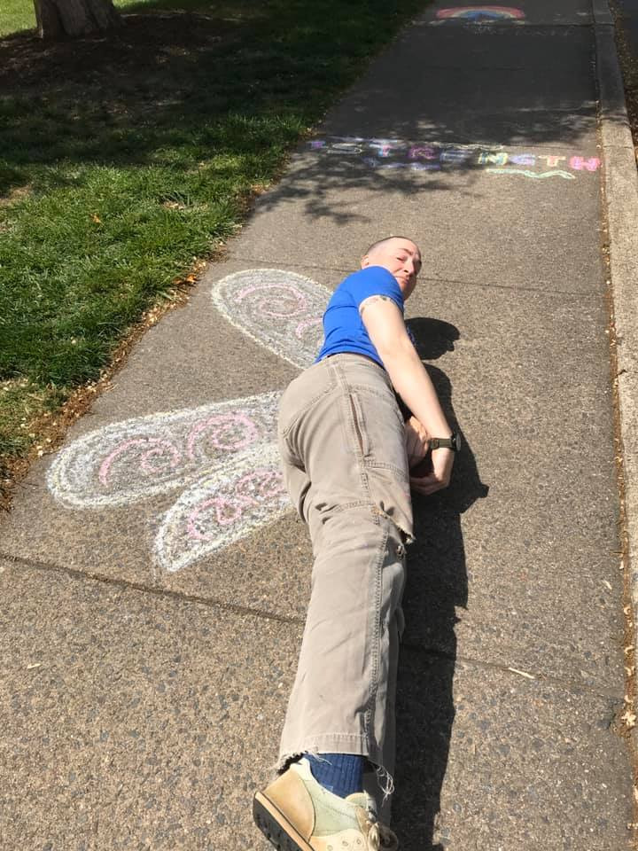 full-grown adult lying on their side on the sidewalk with butterfly wings drawn in chalk behind them.